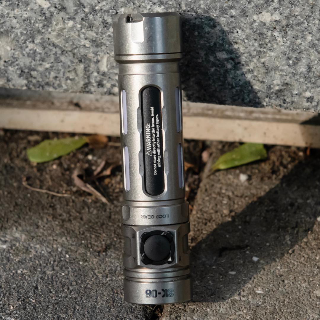 A silver tactical flashlight with a black power button and a black label on the side, photographed standing upright on a gray concrete surface outdoors.