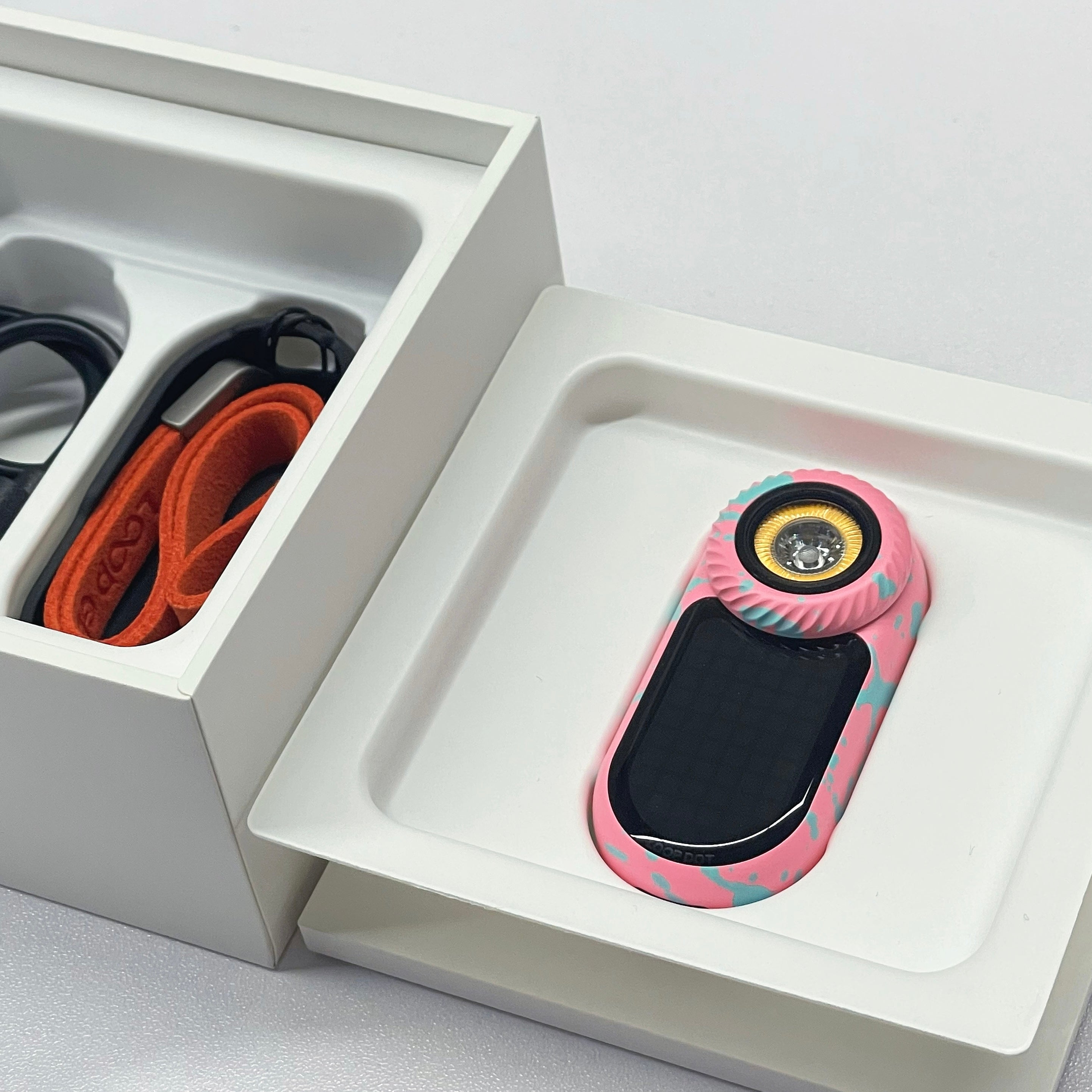 A pink and teal patterned electronic device with a black screen and gold circular detail, displayed in a white tray next to its packaging box.