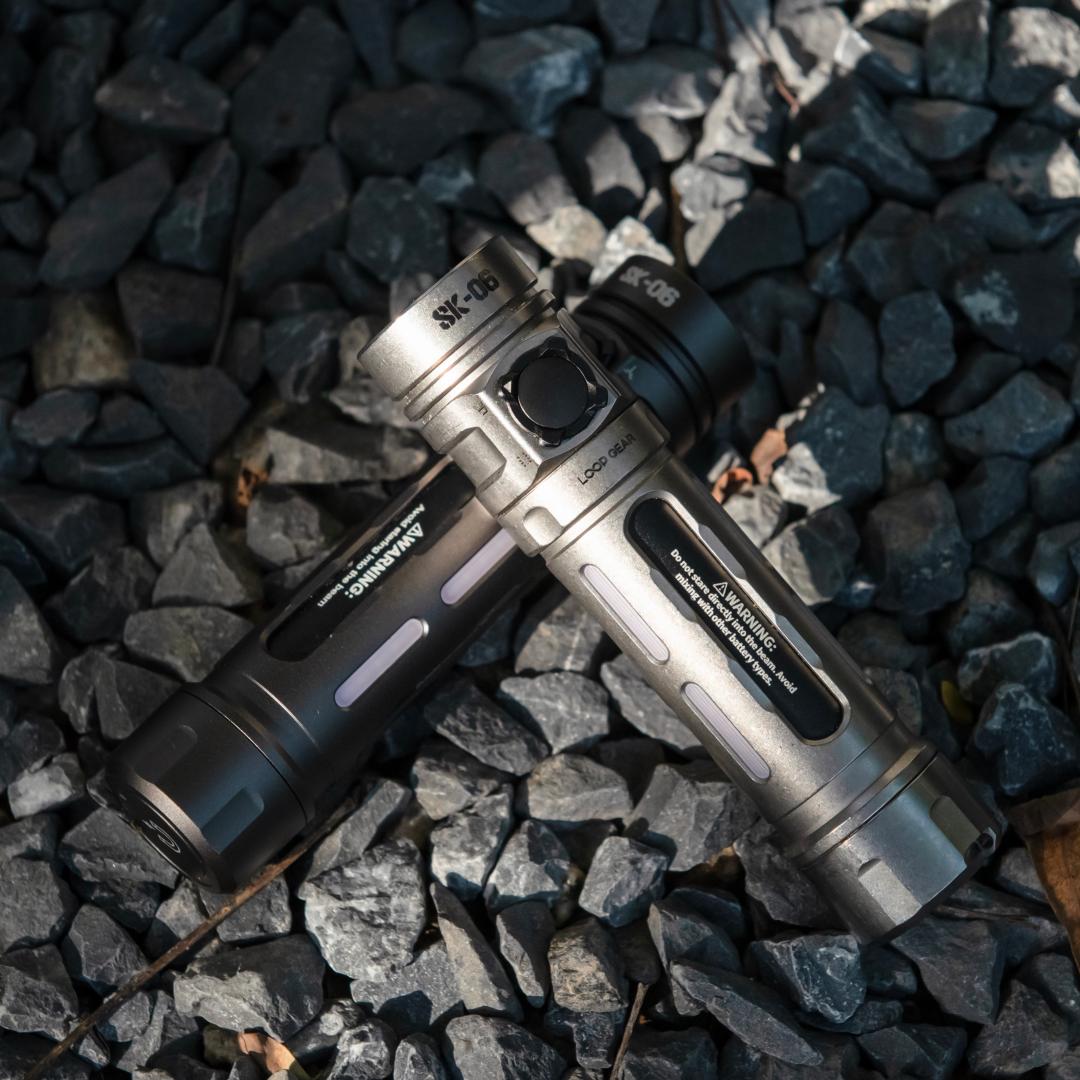 Two tactical LED flashlights, one silver and one black, crossed over each other on a bed of dark gray gravel.